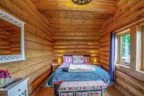 Ein Schlafzimmer in einer Blockhütte mit einem Bett, Nachttischlampen, einem kleinen Tisch mit einer Vase und einem Fenster mit Vorhängen.