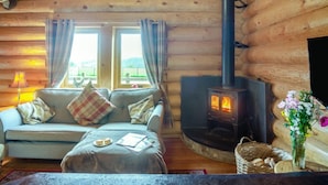 A cozy log cabin interior with a sofa, a table with a plate of food, a fireplace, and a window with curtains. - 2 Bed in Groesffordd Marli (oc-g39079) (Groesffordd Marli)