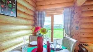 A wooden cabin interior with a dining table set for two, a red vase with flowers, and a framed botanical illustration on the wall. - 2 Bed in Groesffordd Marli (oc-g39079) (Groesffordd Marli)