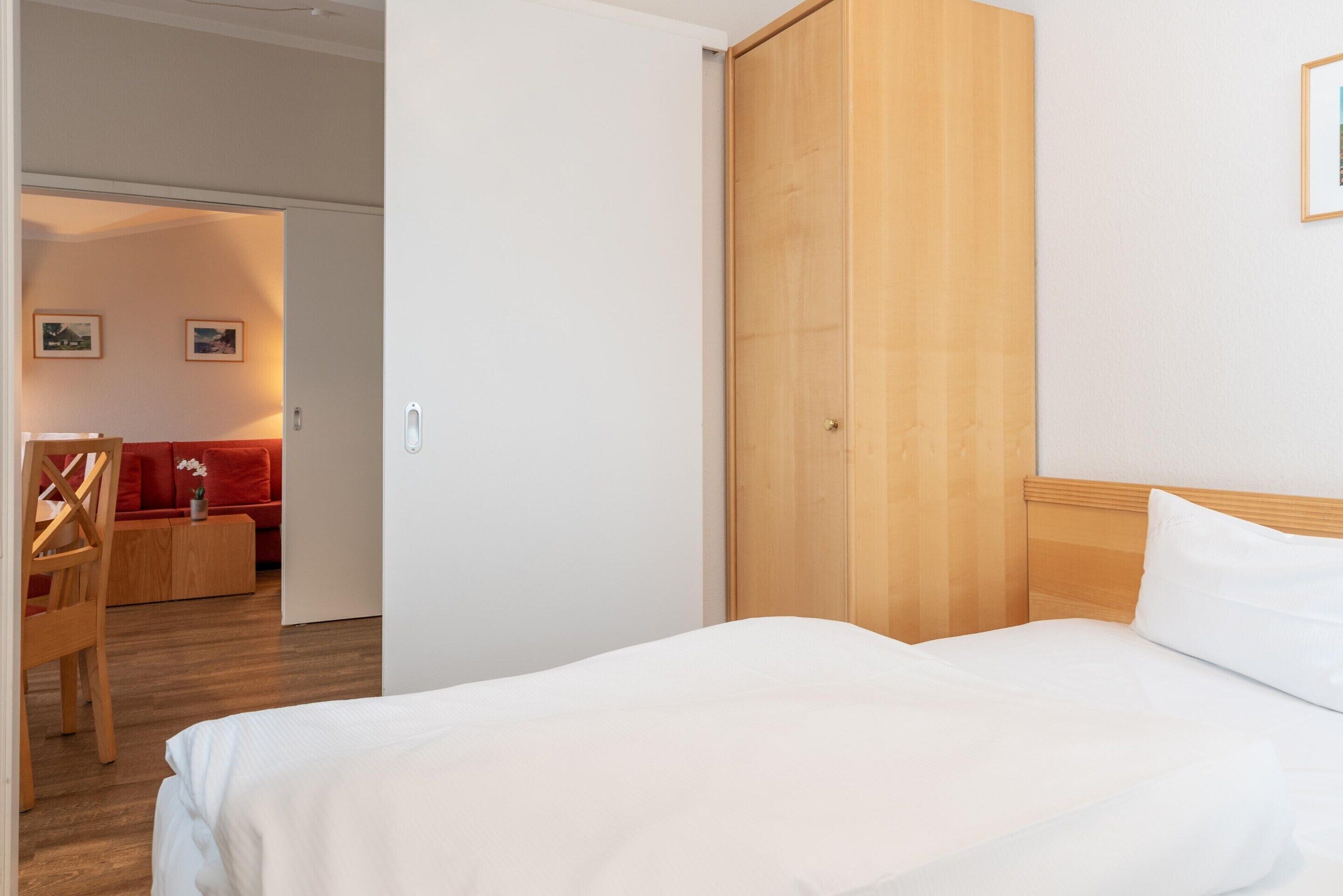 A hotel room with a wooden wardrobe, a bed with white bedding, a wooden floor, a red sofa, and a wooden chair.
