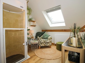 Una habitación con techo inclinado, tragaluz, sillón estampado, una mesita redonda y una cesta de mimbre.