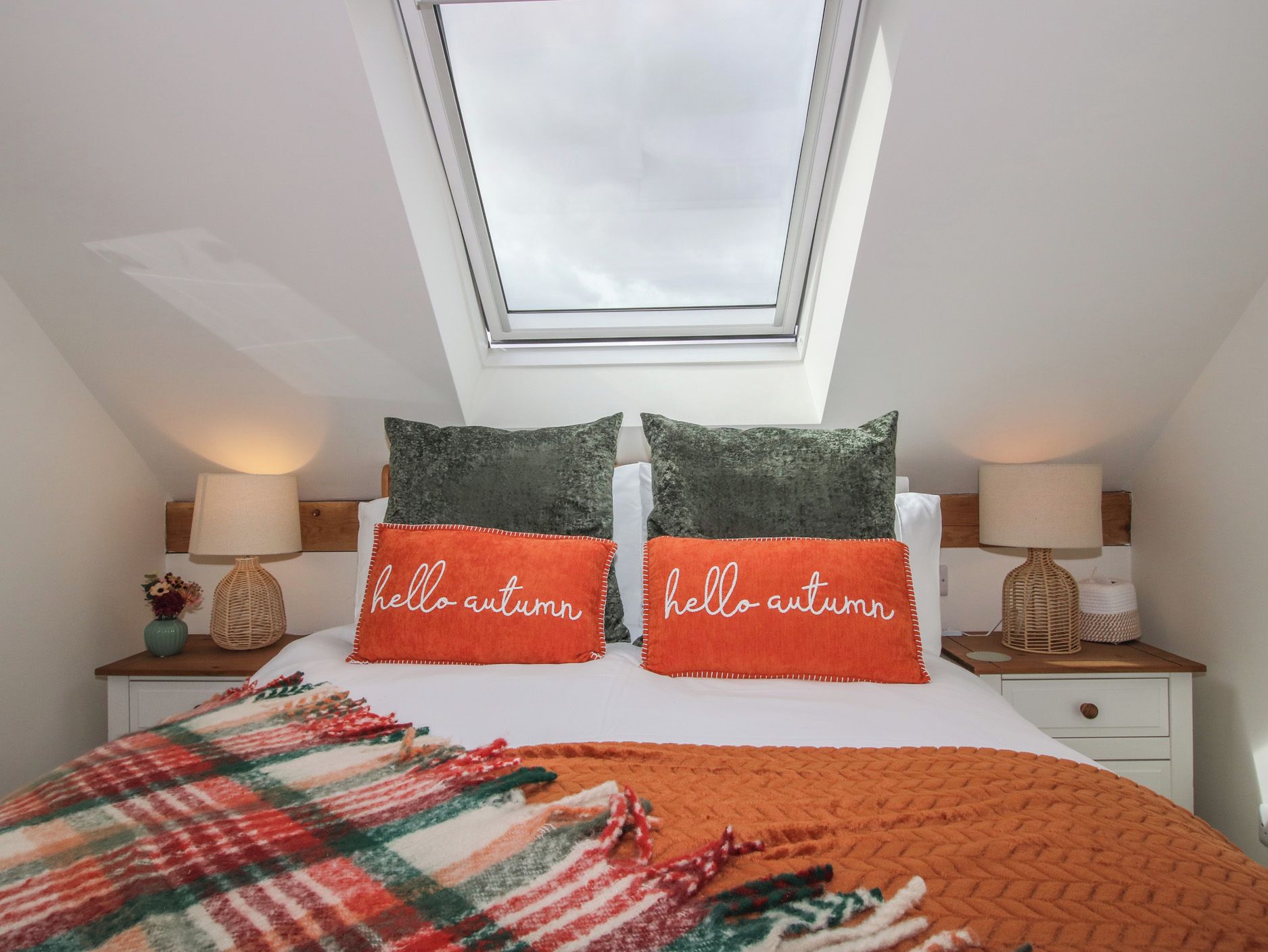 A bedroom with a skylight, two pillows with 'hello autumn' text, a plaid blanket, and bedside tables with lamps.