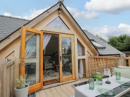 Una cabaña de madera con techo inclinado, puertas de cristal amplias y un pequeño porche con mobiliario exterior.