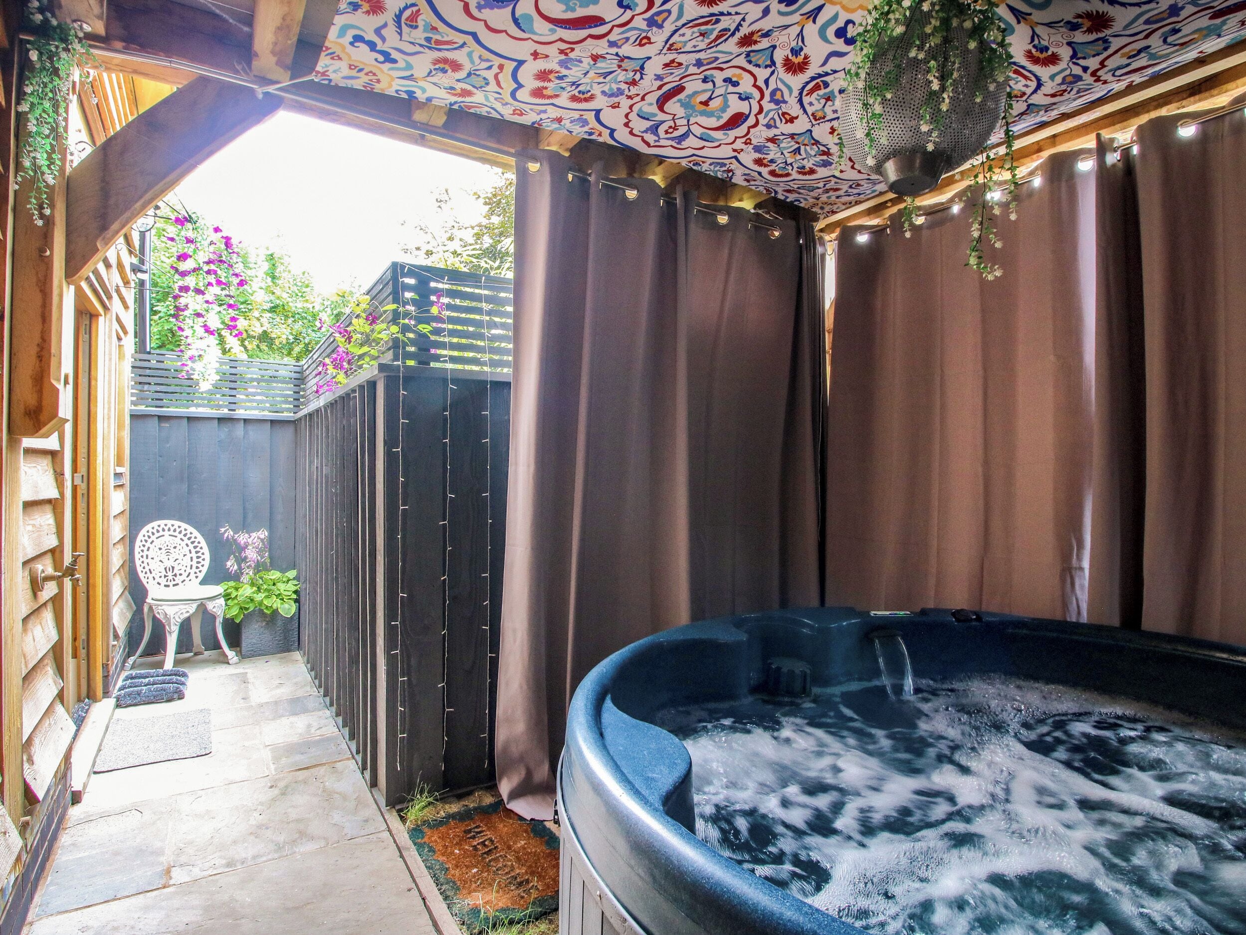 Uma banheira de hidromassagem com um deck de madeira, um espaço de varanda coberto e vista para um jardim com flores roxas.