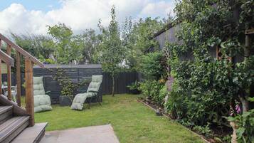 A backyard with a wooden deck, a green cushioned chair, a small garden with various plants, and a wooden fence.