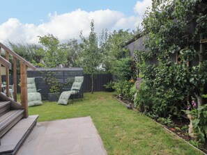 Patio con terraza de madera, silla verde acolchada, pequeño jardín con diversas plantas y cerca de madera.