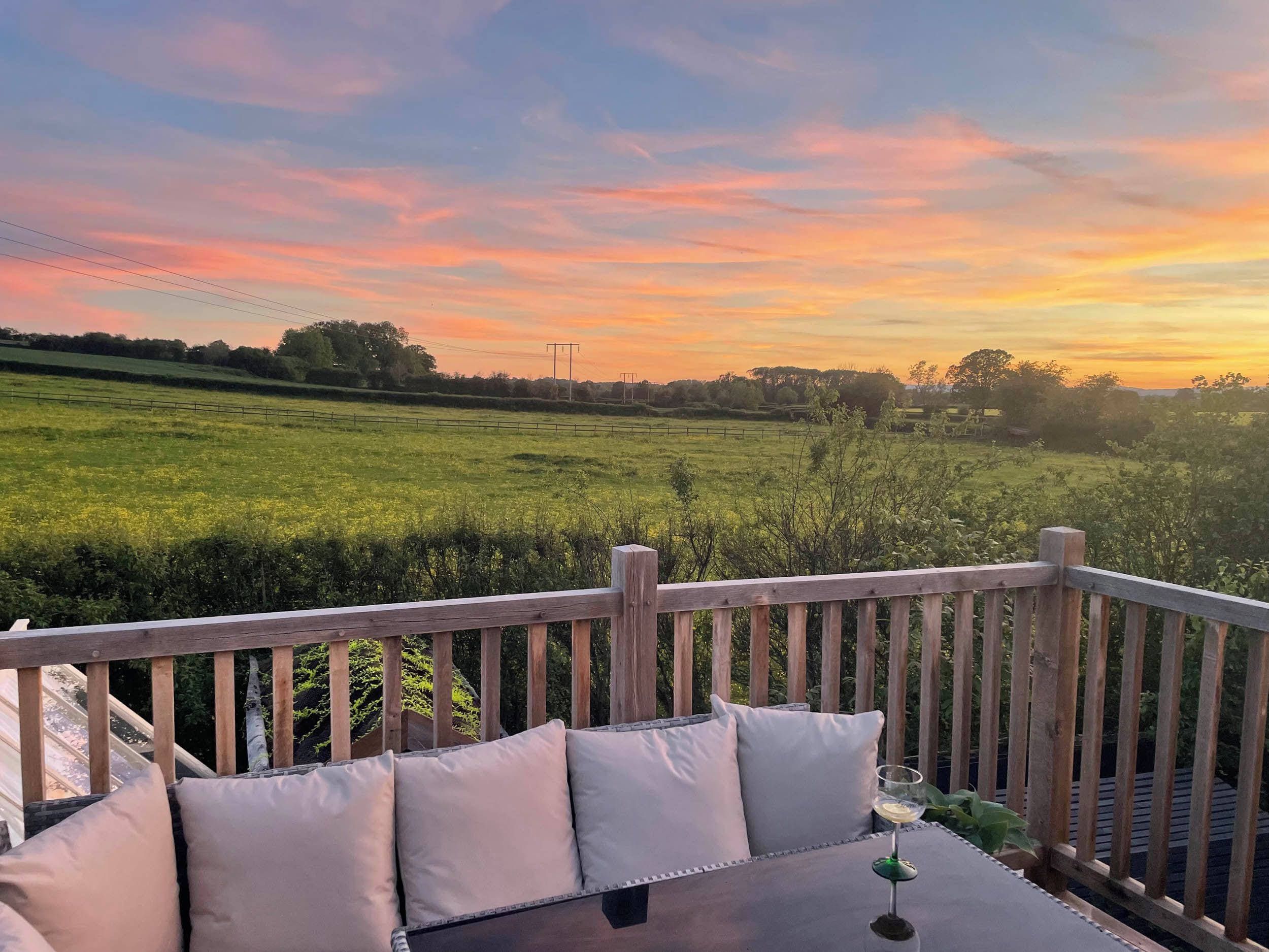 Una terraza de madera con una mesa y cojines, con vistas a un campo verde y una puesta de sol.