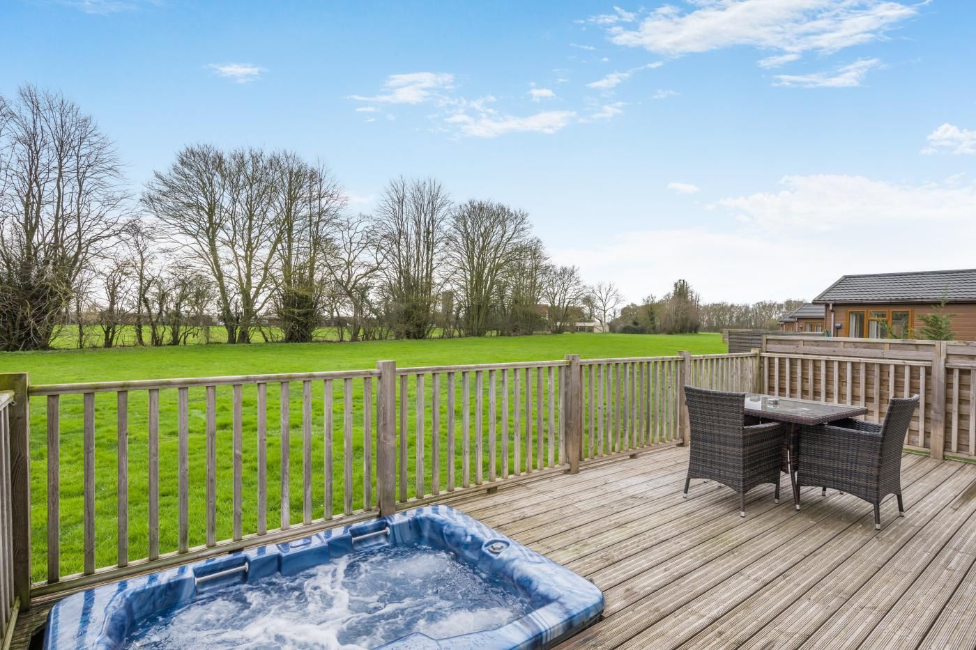 A wooden deck with a hot tub, outdoor furniture, and a view of a green field and trees.