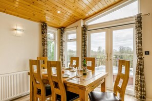 A dining area with a wooden table and chairs, a bottle of wine, and a view of the outdoors through large windows. - 2 Bed in Athelington (oc-h35075) (Athelington)