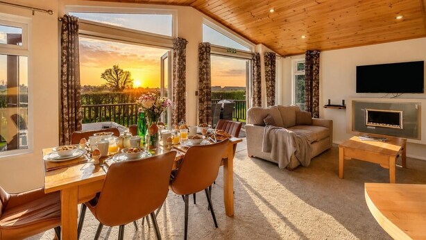Ein Essbereich mit Frühstückstisch, einer Couch und einem Fernseher in einem Raum mit Holzbalkendecke und großen Fenstern.