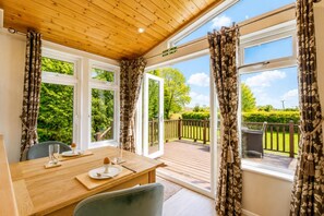 Ein Zimmer mit Holzboden, großen Fenstern mit Vorhängen, einem Holztisch mit Stuhl und Blick auf eine Terrasse und Grünfläche.