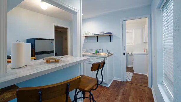 A compact kitchen with a marble countertop, two bar stools, a refrigerator, and a window with blinds.