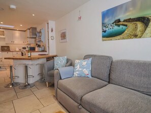 A modern kitchen with white cabinets, a wooden countertop, and bar stools. There is a grey sofa with patterned cushions and a painting of a coastal scene on the wall. - Phoenix Cottage (Lyme Regis)