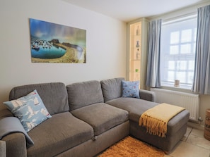 A living room with a grey sofa, a painting of a coastal scene, a window with curtains, and a radiator. - Phoenix Cottage (Lyme Regis)