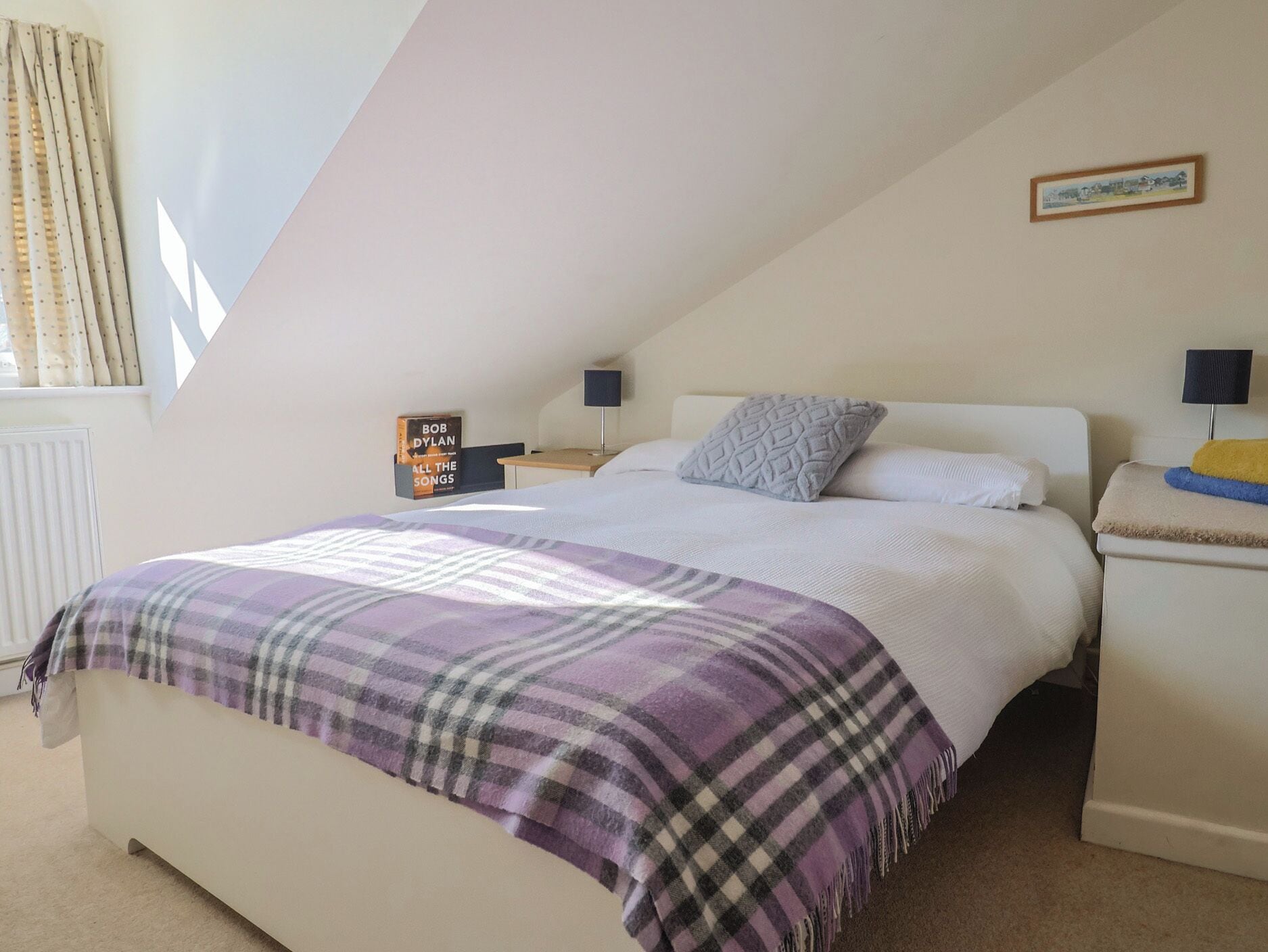 Un dormitorio con cama, mesita de noche, lámpara, un cuadro en la pared y una ventana con cortinas.
