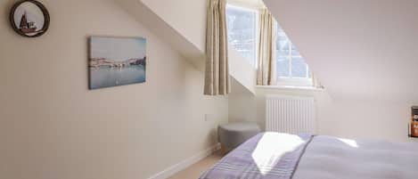 A bedroom with a bed, a window with curtains, a radiator, a small stool, and a painting on the wall.