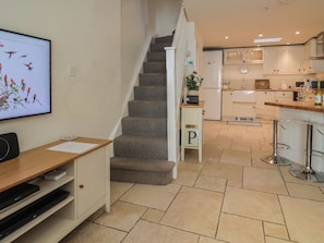 A modern kitchen with a staircase leading to a second floor, a dining area with bar stools, and a flat-screen TV mounted on the wall. - Phoenix Cottage (Lyme Regis)