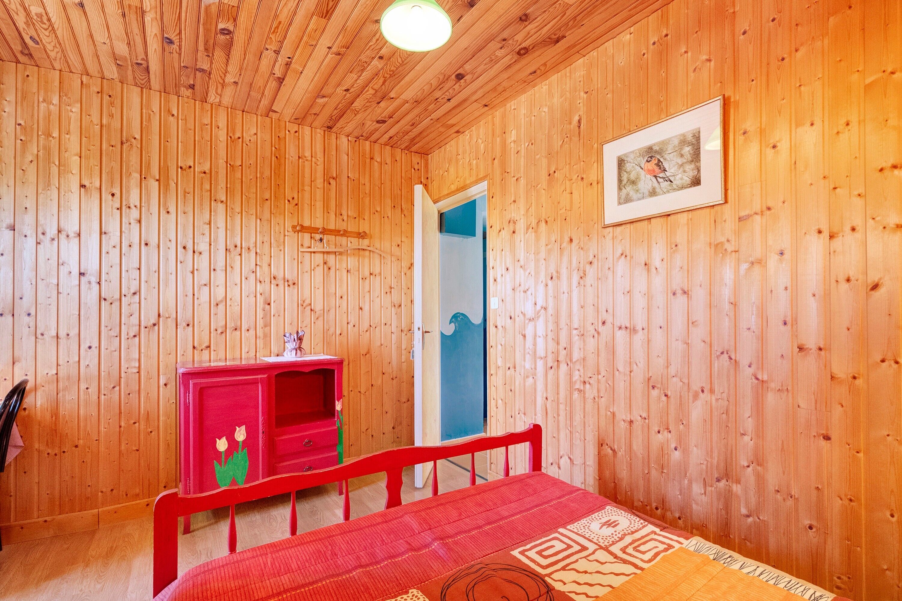 A wooden-paneled room with a bed, a red dresser, and a door leading to another room.