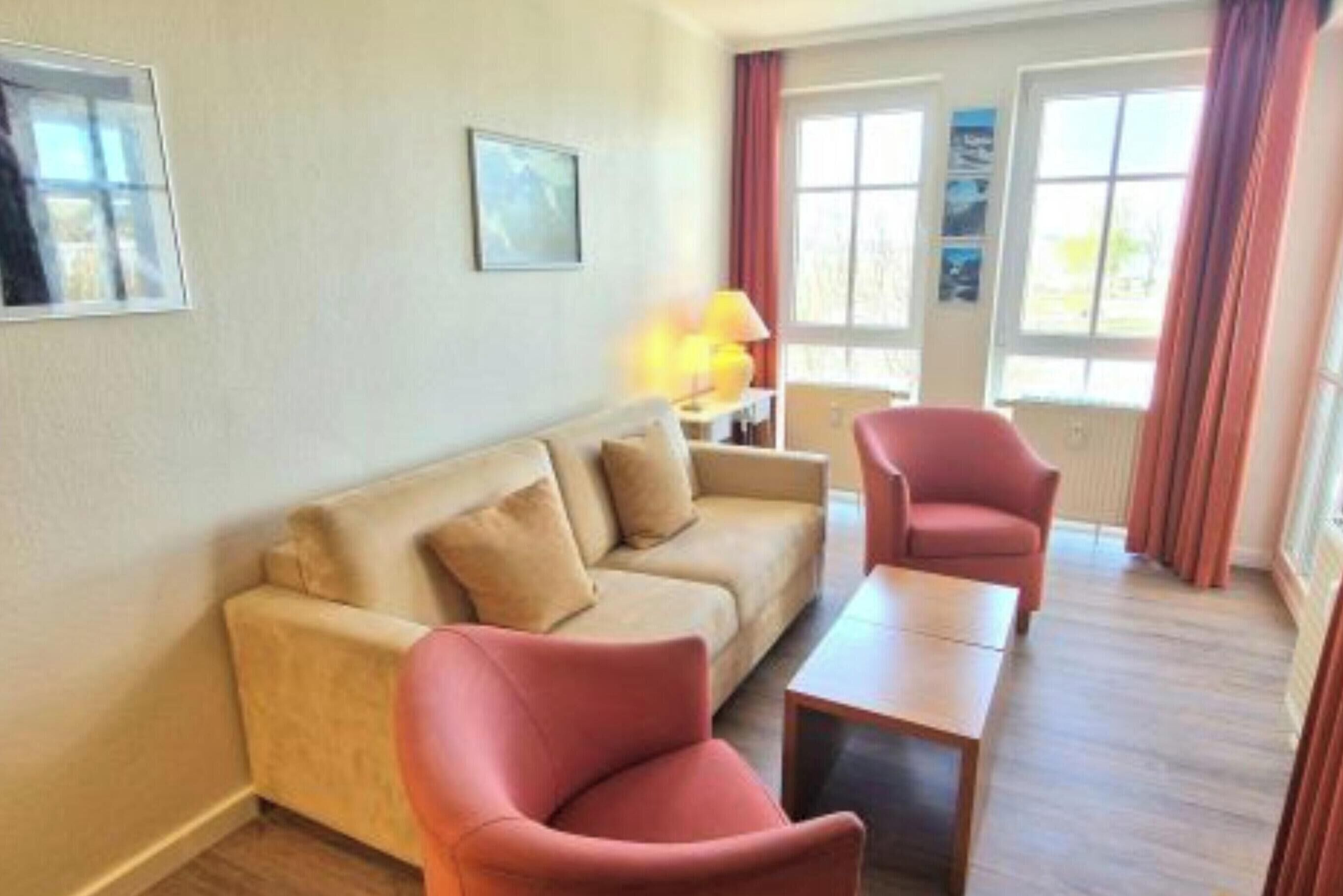 En stue med sofa, lænestol og sofabord. To vinduer med gardiner, en lampe på et sengebord og et indrammet billede på væggen.