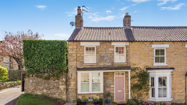 A two-story stone house with a traditional design, featuring a front door, two windows, and a small porch area with potted plants. - 2 Bed in Ampleforth (oc-c37505) (Ampleforth)