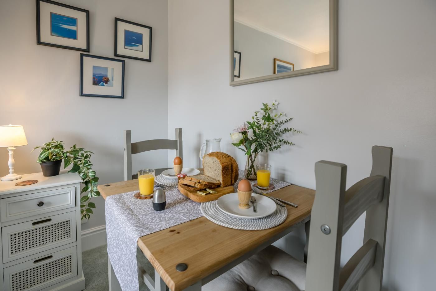 A dining area with a wooden table, chairs, a lamp, a potted plant, and a breakfast setting including bread, eggs, and juice.