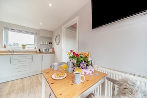 A modern kitchen with a wooden table set for breakfast, a TV mounted on the wall, and a window with a view. - 1 Bed in Gatehouse Of Fleet (oc-h39501) (Gatehouse of Fleet)