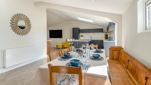 Un espace repas avec une longue table dressée pour six personnes, une cuisine en arrière-plan et un design moderne au niveau du plafond.