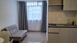 A modern living room with a sofa, a dining table, and chairs, a kitchen with cabinets and a sink, and a large window with curtains. - Comfortable 1-bedroom apartment #301 near the U.S. Embassy (Bogotá, D.C.)