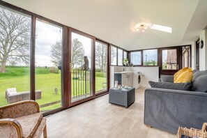 A spacious living area with a large sliding glass door leading to a garden, a grey sofa, a small table with decorative items, and a washing machine in the background. - 4 Bed in Snape (oc-t36355) (Snape)