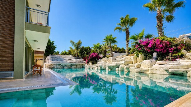 A clear swimming pool with a stone wall and palm trees, a wooden deck, and a building with a balcony. - SARUM ÇEŞME (İzmir)