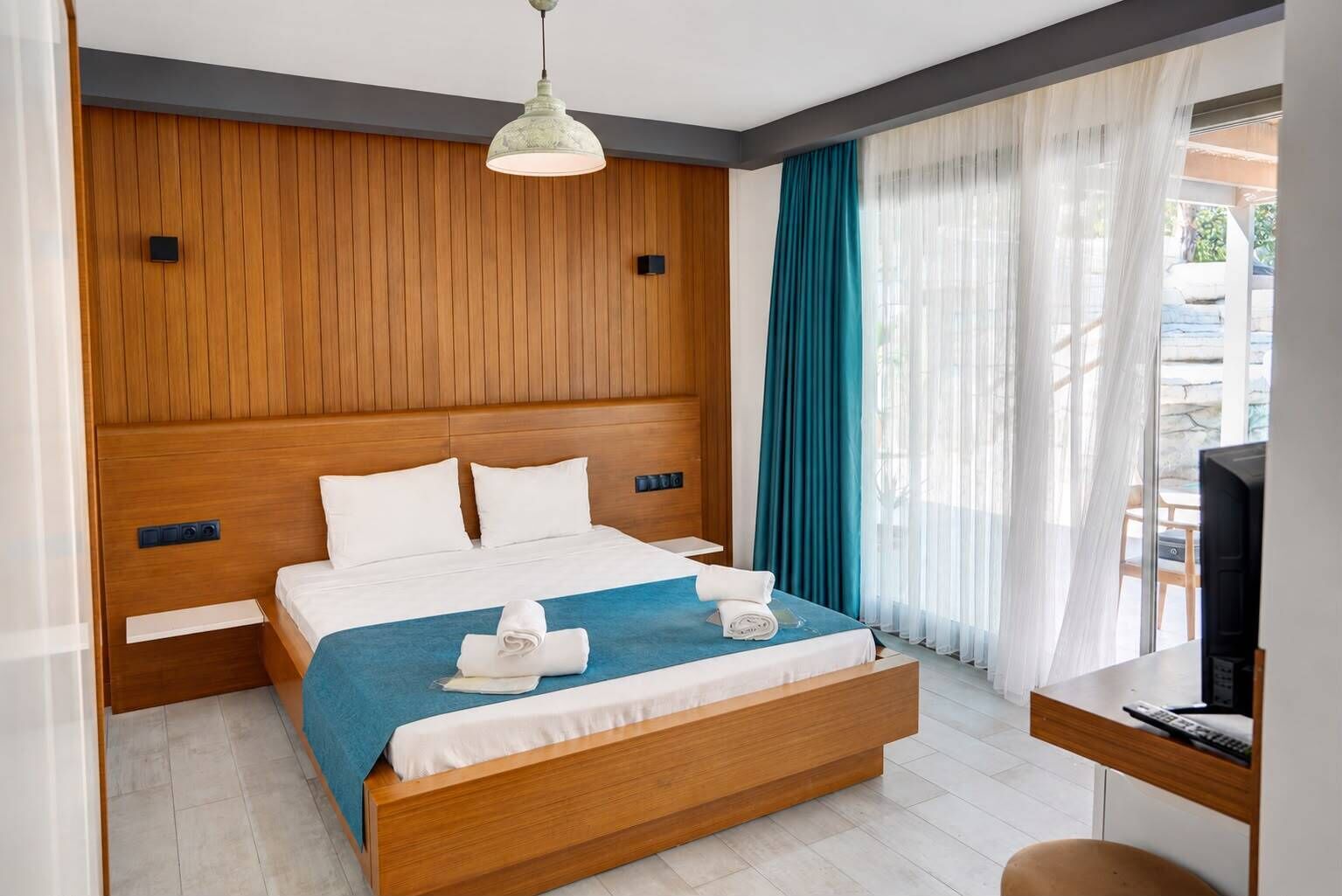 A hotel room with a wooden headboard, a bed with white bedding, a blue throw blanket, two rolled towels, a television on a wooden table, and a window with teal curtains.