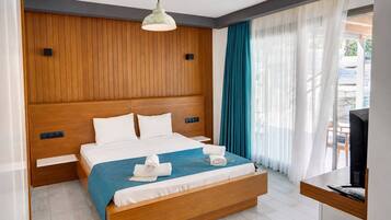 A hotel room with a wooden headboard, a bed with white bedding, a blue throw blanket, two rolled towels, a television on a wooden table, and a window with teal curtains.
