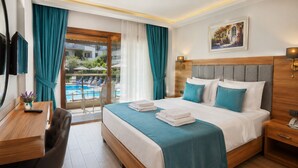 A hotel room with a bed, a desk, a chair, a bedside table, a lamp, and a view of a swimming pool. - SARUM ÇEŞME (İzmir)
