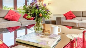 Una sala de estar con una mesa de centro de vidrio, un libro titulado 'The Elements of Organic Gardening', un jarrón con flores y dos sofás grises con cojines rojos.