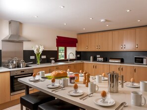 A modern kitchen with wooden cabinets, stainless steel appliances, and a central island with breakfast items. - 4 Bed in Llandovery (oc-bn208) (Llanfair-ar-y-Bryn)
