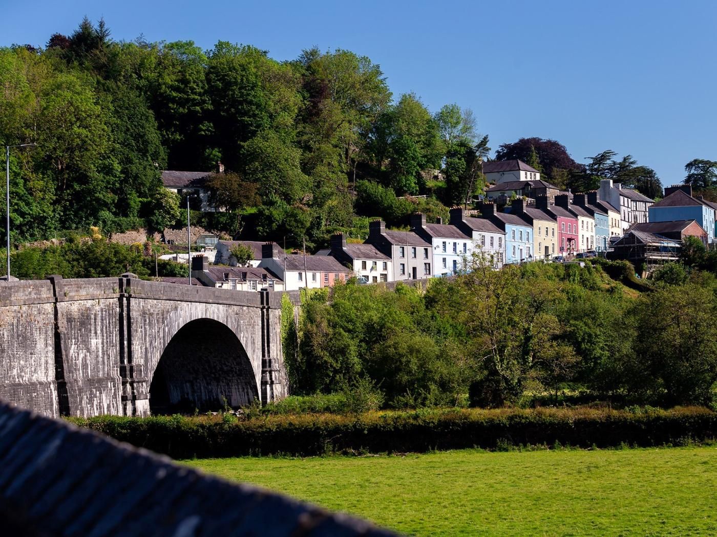 Eine Steinhöhenbrücke mit Bogen über einer grünen Wiese, die zu einer Reihe farbenfroher Häuser an einem Hang führt.