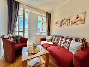 Ein gemütliches Wohnzimmer mit einem roten Sofa, einem roten Sessel, einem Glastisch und Blick auf die Stadt durch große Fenster.