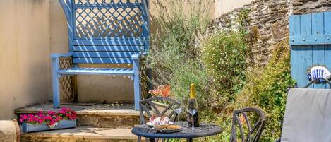 A patio with a blue chair, a round table with a bottle and glasses, and a small flower arrangement.