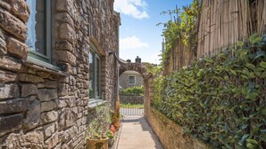 A narrow stone pathway leading to a traditional stone building with a wooden fence and lush greenery on one side. - 3 Bed in Plymouth (oc-49520) (Newton Ferrers)