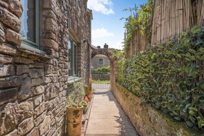 A narrow stone pathway leading to a traditional stone building with a wooden fence and lush greenery on one side. - 3 Bed in Plymouth (oc-49520) (Newton Ferrers)