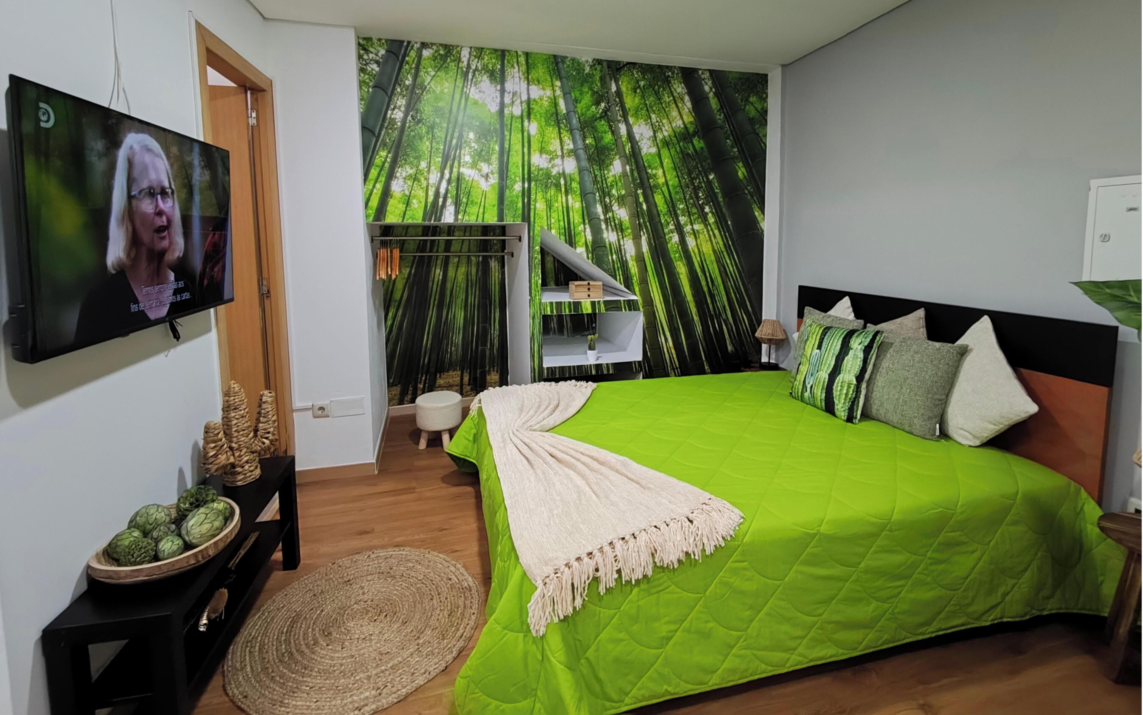 Una camera da letto con copripiumino verde, televisione montata a parete e un murale raffigurante una foresta di bambù.