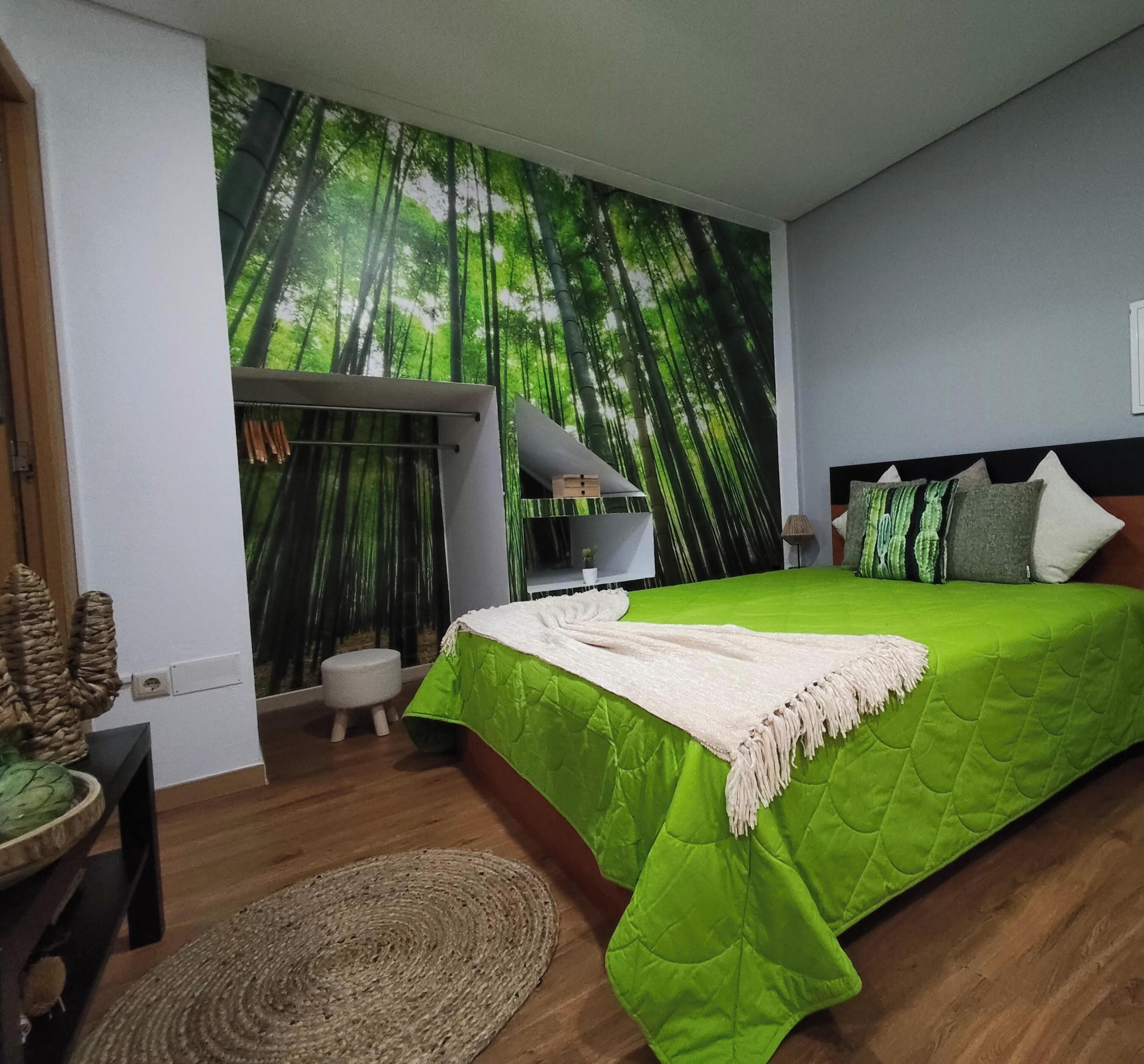 Slaapkamer met een groene sprei, een witte plaid, een klein krukje, een geweven vloerkleed en een muur met een bosreliëf.