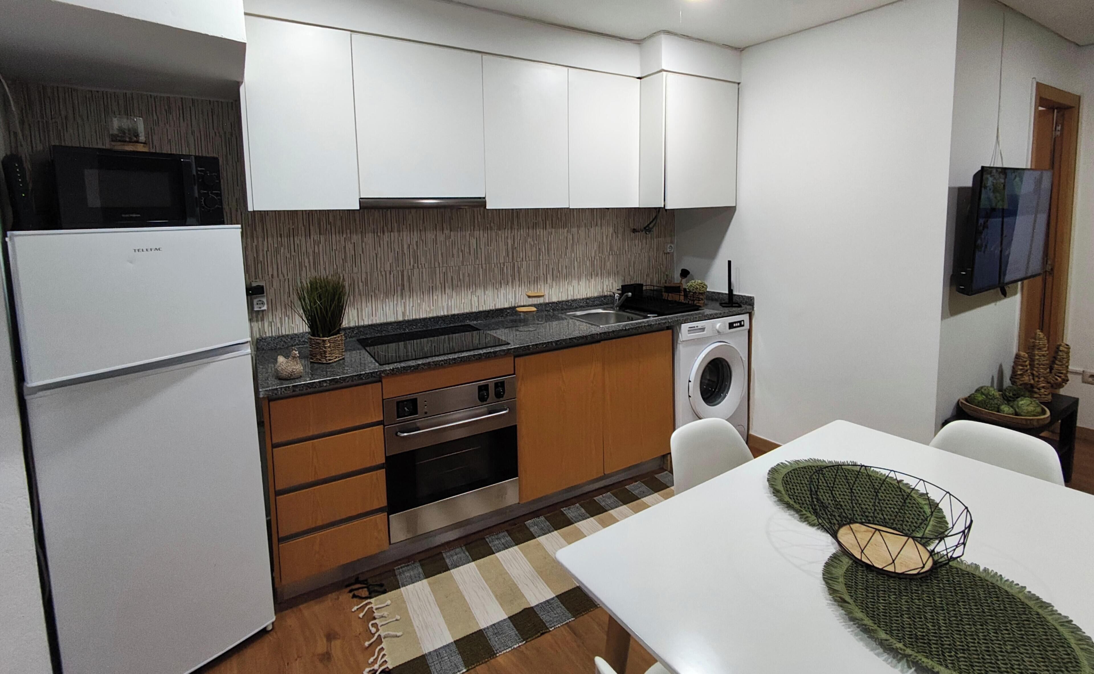 A compact kitchen with white cabinets, a built-in microwave, a washing machine, and a dining area with a table and chairs.