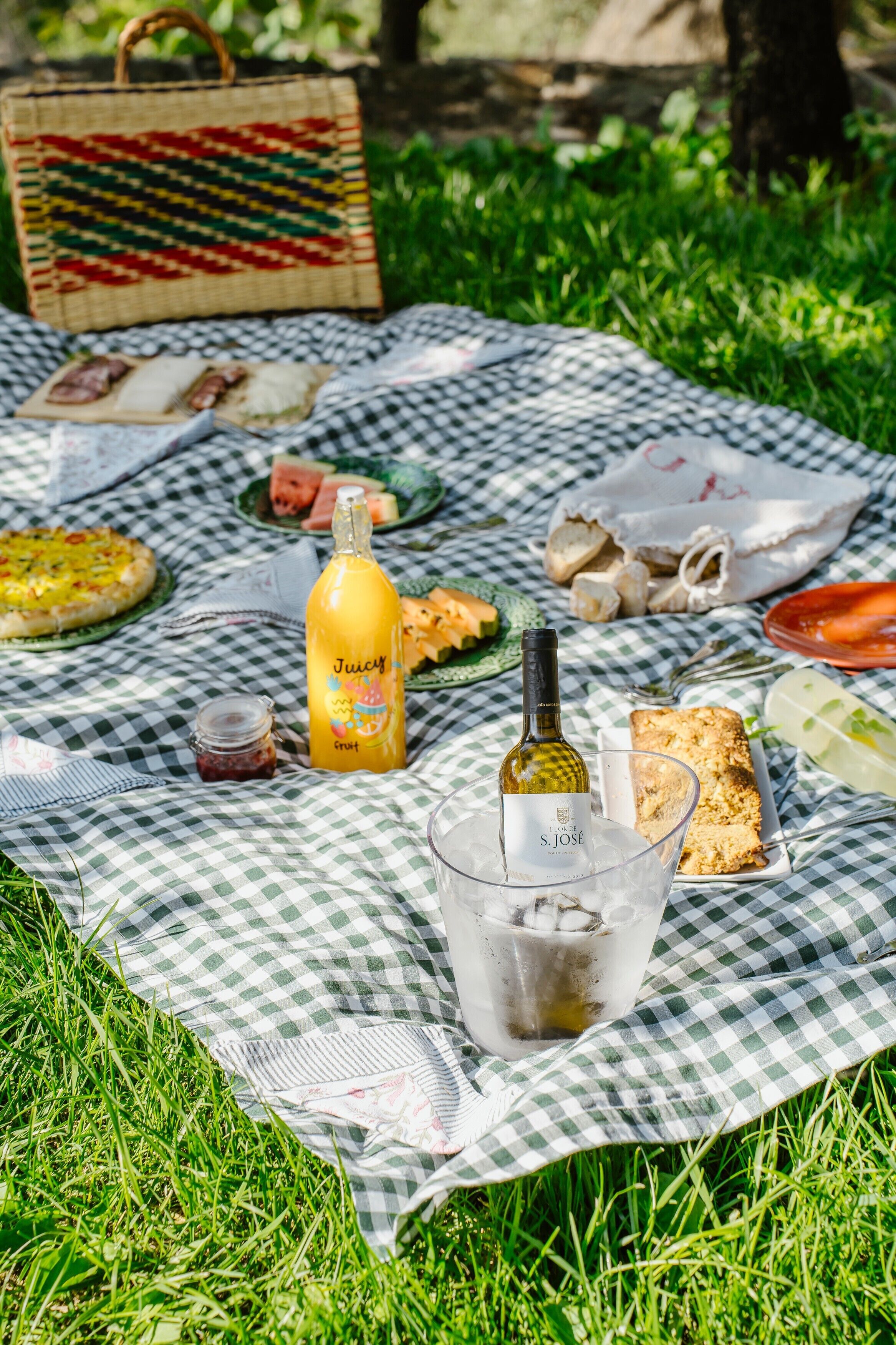 A picnic setup on a checkered blanket with food, a bottle of juice, and a glass of water.