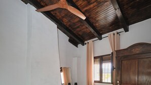 A bedroom with a bed, a wooden wardrobe, a desk with a chair, a window with curtains, and a ceiling fan.