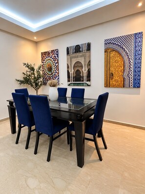 Un espace repas comprenant une table à plateau en verre, six chaises bleues et trois œuvres d’art accrochées au mur.