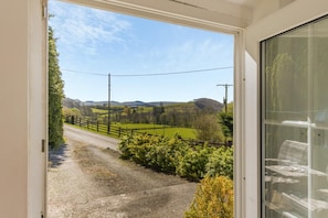 Vista desde una habitación con puerta que da a un camino rural, rodeado de vegetación y colinas.
