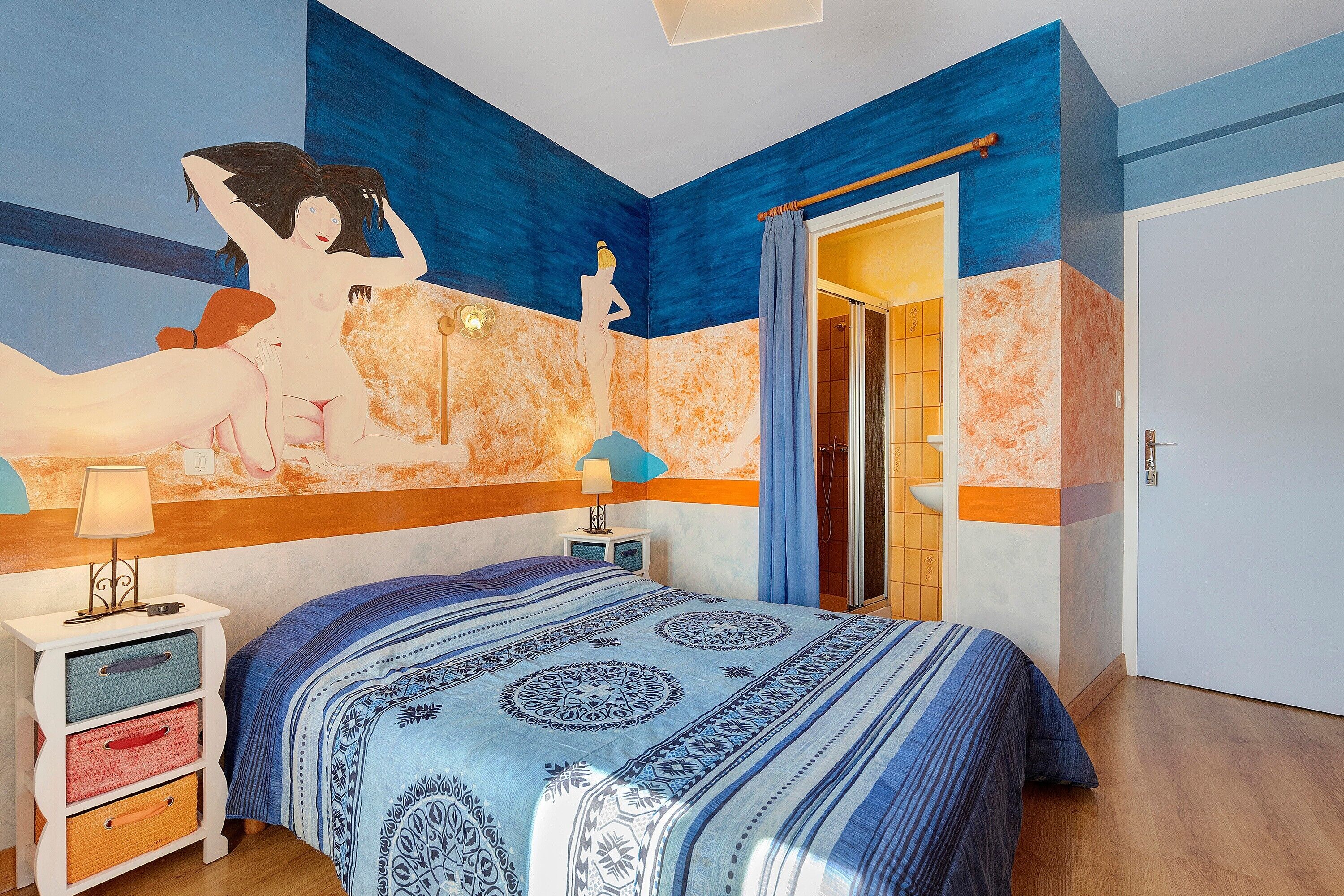 A bedroom with a bed, a nightstand, a wall with a mural, a door, and a shower area.