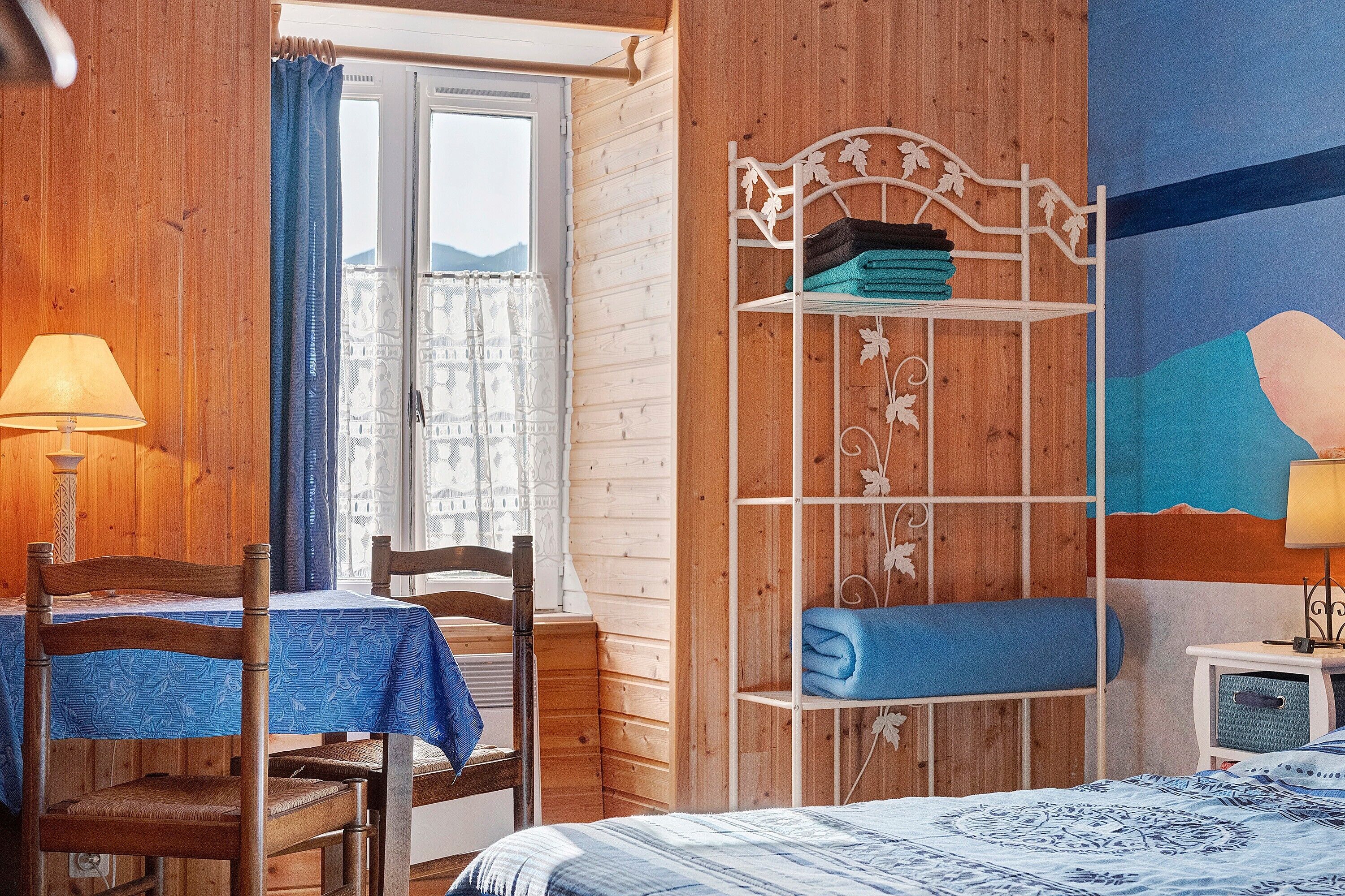 Een kamer met een bed, een stoel, een klein tafeltje, een plank met opgevouwen handdoeken en een raam met gordijnen.