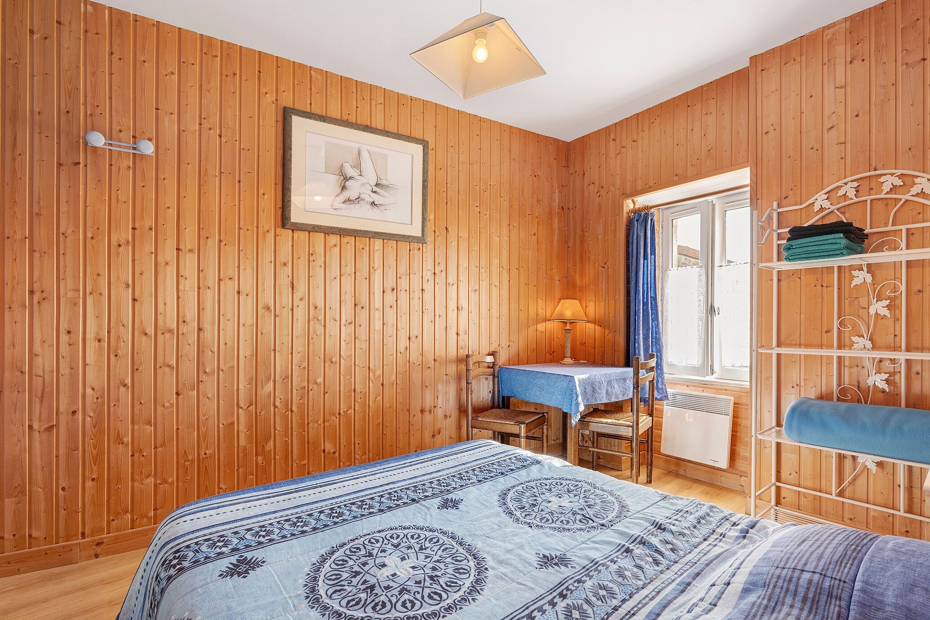 Ein Zimmer mit Holzwänden, einem Bett, einem Tisch, einem Stuhl und einem mit Vorhängen drapierten Fenster.